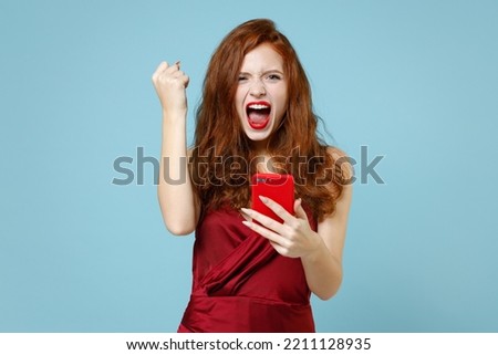 Young caucasian fun excited successful lucky readhead curly woman in red party dress hold mobile cell phone do winner gesture clench fist celebrating isolated on pastel blue background studio portrait
