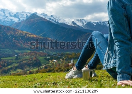 Close-up of a woman in jeans with a teapot and a cup of coffee sitting on the edge and watching the amazing beautiful mountain landscape on an autumn day.Concept of travel, outdoor activities