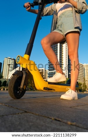Close-up of slender tanned female legs in denim shorts, sneakers are standing on an electric scooter. Young woman poses with her yellow electric eco scooter in a city park. Vertical photo