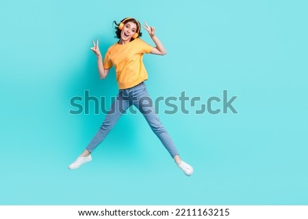 Full length photo of adorable sweet girl dressed yellow t-shirt enjoying songs jumping high showing v-signs isolated teal color background