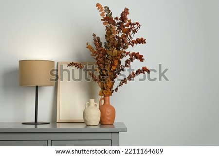 Stylish vases, dried eucalyptus branches and table lamp on chest of drawers near white wall indoors, space for text. Interior design
