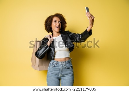 happy african american woman in stylish leather jacket holding shopping bags and taking selfie on yellow
