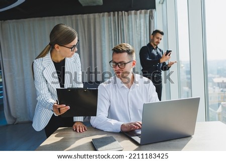 Group of young business colleagues in classic clothes sitting at table with laptop and documents, discussing details, looking at each other. The atmosphere of a working day in the office