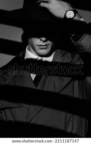 A dark silhouette of a dangerous man in a hat. The style of film noir. Private detective, spy, investigation concept. Dramatic light about the blinds.