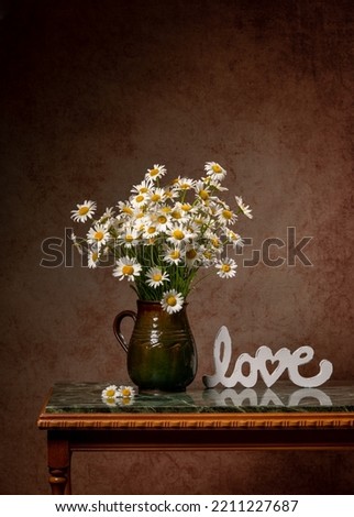 A large bouquet of white daisies in a brown clay jug on a marble green table. Near the glass of white wine and the inscription love. Fun summer still life. Thoughtfulness