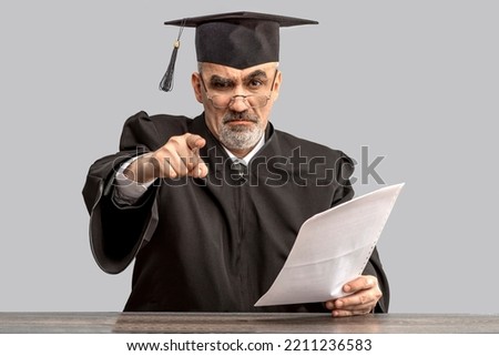 Catch in plagiarism concept. Angry Academician, Professor, Prof is wearing a cap with Tassel and black gown pointing finger at the camera. Senior educator learned man from a university or college. Royalty-Free Stock Photo #2211236583