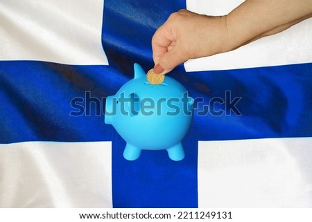 Hand of elderly woman putting coin into piggy bank on Finland flag background. Hand putting coin to piggy bank. Retirement savings, piggy bank. Concept saving money and retirement fund in Finland