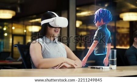 Young asian woman wearing VR headset conversing with a cartoon character avatar via an futuristic screen hologram. Futuristic communication scifi concept. 3D rendering picture. Augmented reality. Royalty-Free Stock Photo #2211249845