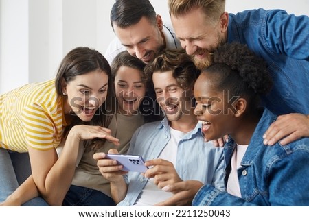 Man uses mobile phone to show vacation photos to his friends during meeting at home. Cheerful multiracial millennial people sitting together on sofa and smiling happily looking at smartphone screen.