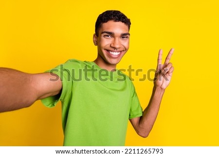 Closeup selfie photo of young positive smiling guy wear green t-shirt good mood showing v-sign hello world blogger online isolated on yellow color background