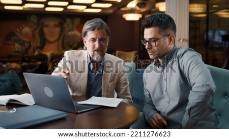 Mature man using laptop, talking with serious businessman, showing papers, documents. Business meeting in office restaurant. Signing contract for new economic strategy of company