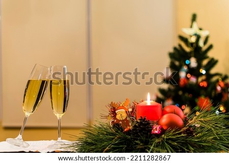 Candle with Christmas objects around and next to two champagne glasses and a Christmas tree in the background.