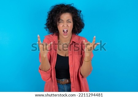 Born to rock this world. Joyful brunette arab woman wearing pink shirt over blue background screaming out loud and showing with raised arms horns or rock gesture.
