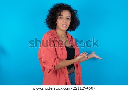 brunette arab woman wearing pink shirt over blue background Inviting to enter smiling natural with open hands. Welcome sign.