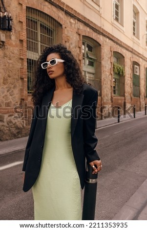 Fasionable young african girl in stylish sunglasses looking into distance stands on europe street. Brunette with curly hair wears coctail dress and black jacket. Concept fasion, female beauty. Royalty-Free Stock Photo #2211353935