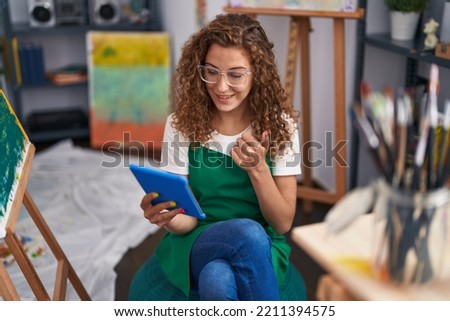 Young caucasian woman painter sitting at art studio doing video call with tablet smiling happy and positive, thumb up doing excellent and approval sign 