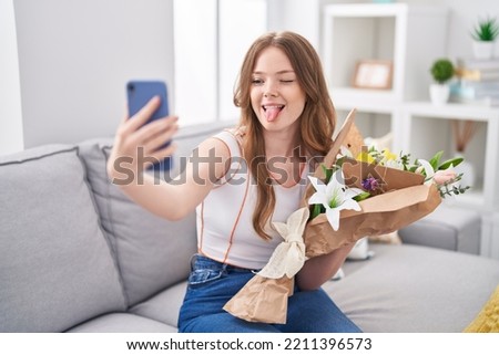 Caucasian woman holding bouquet of white flowers taking a selfie picture sticking tongue out happy with funny expression. 