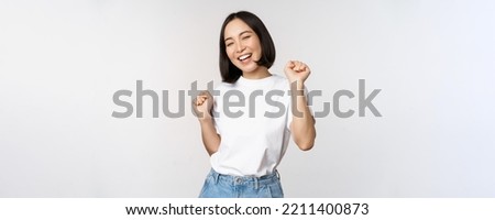 Happy dancing korean girl posing against white background, wearing tshirt Royalty-Free Stock Photo #2211400873