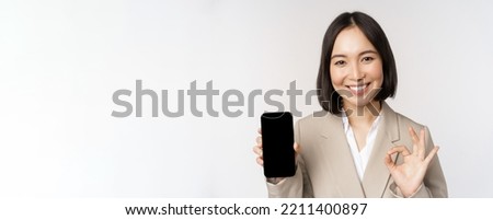 Smiling asian woman showing smartphone screen and okay sign. Corporate person demonstrates mobile phone app interface, standing over white background