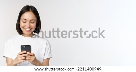 Portrait of smiling asian woman using mobile phone, chatting, texting message, standing in tshirt over white background