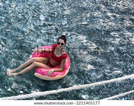 Young woman in bikini swims on an inflatable donut lap in black sea with clear water overlooking the rocks. The girl is smiling, enjoying herself and looking at the camera.