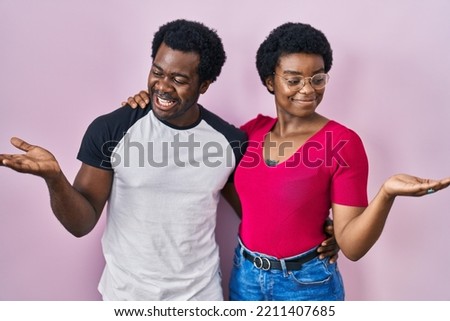 Young african american couple standing over pink background smiling showing both hands open palms, presenting and advertising comparison and balance 