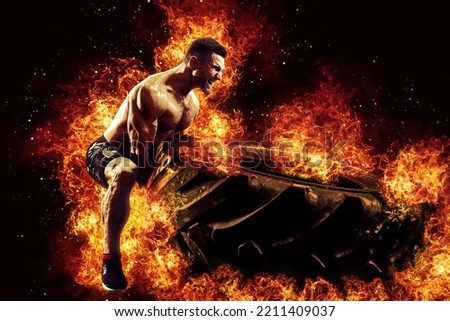 Muscular fitness shirtless man moving large tire in flames, concept lifting, workout cross training.  Royalty-Free Stock Photo #2211409037
