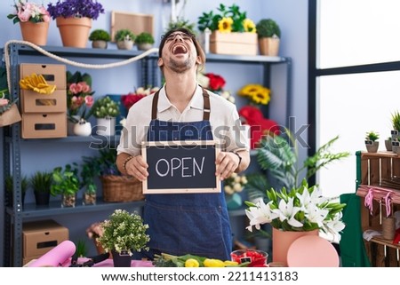 Hispanic man with long hair working at florist holding open sign angry and mad screaming frustrated and furious, shouting with anger looking up. 