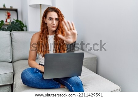 Young redhead woman using laptop at home with open hand doing stop sign with serious and confident expression, defense gesture 