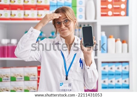 Young caucasian woman working at pharmacy drugstore showing smartphone screen stressed and frustrated with hand on head, surprised and angry face  Royalty-Free Stock Photo #2211431053