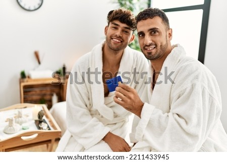 Two hispanic men couple holding credit card sitting on massage table at beauty center
