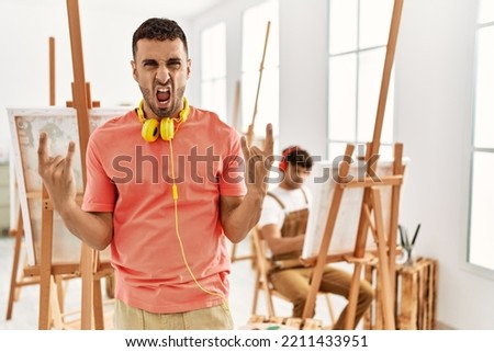 Young hispanic man at art studio shouting with crazy expression doing rock symbol with hands up. music star. heavy concept. 