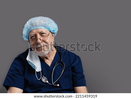 An elderly doctor with a stethoscope, a cap, a mask and glasses on a gray background, looks kindly into the camera, close-up, space for text