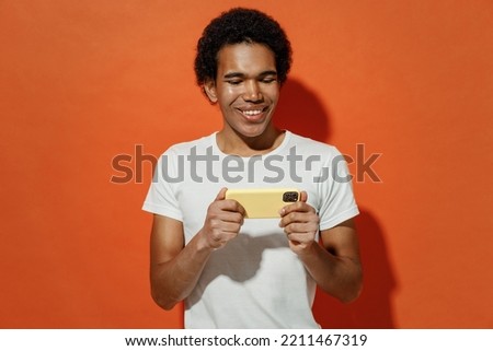 Gambling young black curly man 20s wears white t-shirt using play racing app on mobile cell phone hold gadget smartphone for pc video games isolated on plain pastel orange background studio portrait