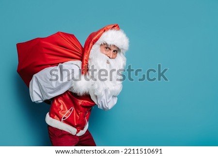 slinking santa claus carrying huge bag of gifts on blue background while looking at camera. New Year concept