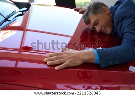 Middle-aged man in car dealership hugging hood of red car