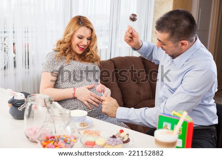 Picture of a young pregnant woman and her husband playing drums with lollipops on her belly, sitting at the kitchen table full of sweets