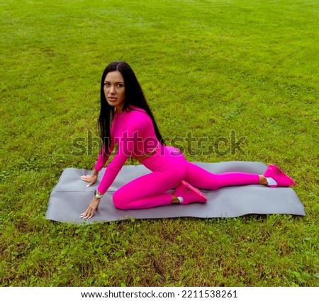 Young athletic brunette woman in pink clothes doing stretching on a sports mat. A woman with long hair is exercising in the park on the green grass. Portrait of a beautiful sports woman