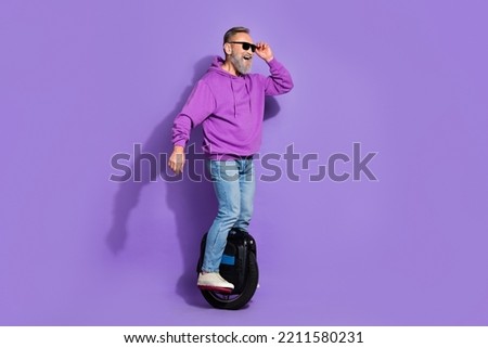 Portrait of senior funky optimistic cheerful man wear purple hoodie driving monoscooter hold sunglass isolated on violet color background Royalty-Free Stock Photo #2211580231