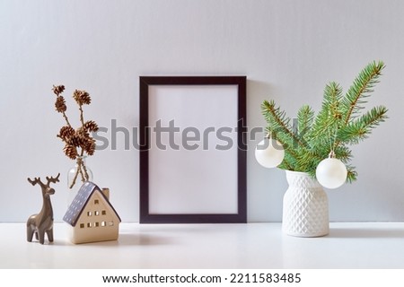 Mock up black poster frame with christmas decoration in home interior, scandinavian style. Christmas little house and deer, green fir branches in a vase on a white table