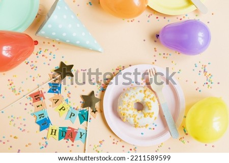Tasty donut on a pink plate near colorful balloons, birthday hat, happy birthday garland on a beige background with confetti. Happy birthday party table setting concept with copy space