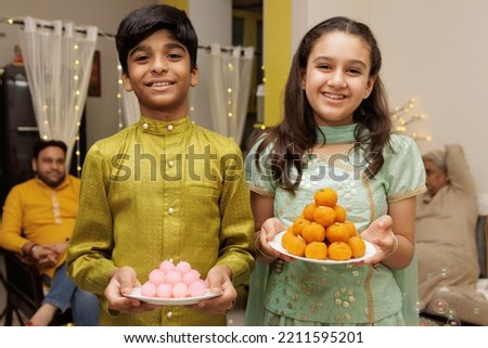 Young girl and boy, kid, dressed up in ethnic wear holding plate of sweets with smile expression pose for photo with family in background celebrating diwali Hindu festival Laxmi poojan