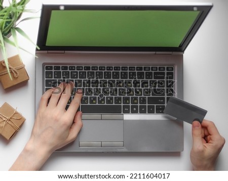 Hands of a young caucasian man holding a bank card in his hand and typing on the keyboard of a laptop with a green screen, which lies on a white desktop with small craft gift boxes and a flower in a