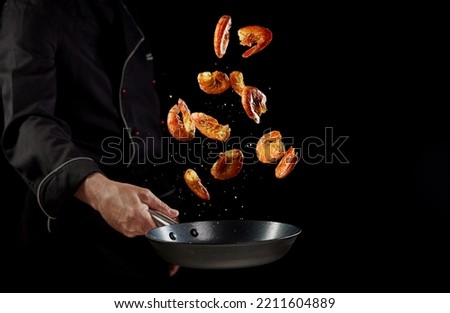 Crop anonymous male chef tossing prawns in frying pan while cooking delicious dinner with seafood against black background Royalty-Free Stock Photo #2211604889