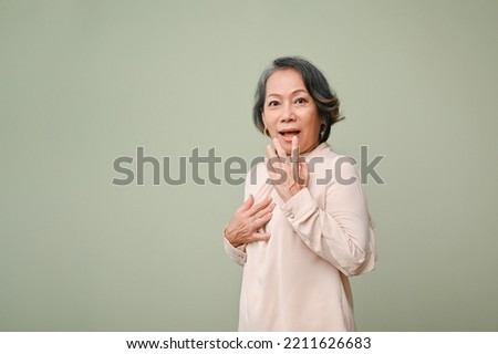 Surprised and excited 60s senior Asian woman in casual standing against isolated green studio background. shocked pose for advertising your big shopping deal.