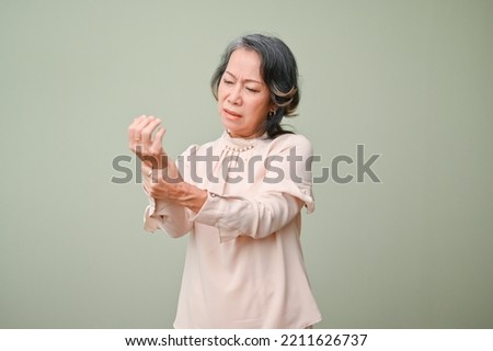 Unhappy and distressed elderly Asian woman holding her wrist, suffering from severe pain in her hand, massaging her aching wrist, standing against a green isolated background.  Royalty-Free Stock Photo #2211626737
