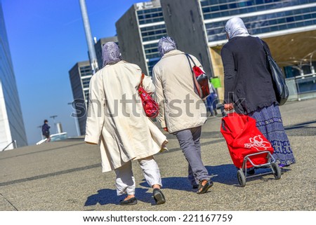 Three women in headscarfs walking in the street with one of them pulling a large red bag on wheels