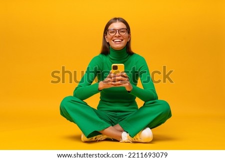 Pretty millennial woman having fun laughing out loud sitting on floor with phone on yellow background browsing social media posts, laughing at funny memes, videos, stories ad reels Royalty-Free Stock Photo #2211693079