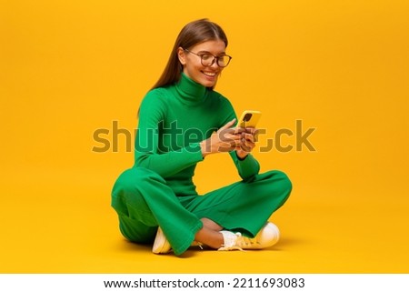 Side view portrait of happy young woman in green clothes sitting on floor on yellow background reading e-book in smartphone app, wearing stylish glasses. People and technology