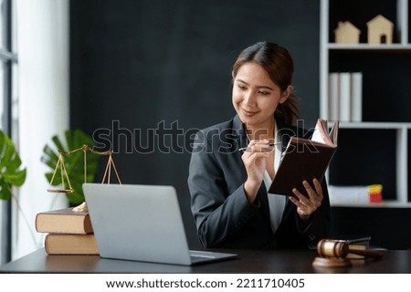 Beautiful Asian woman lawyer sitting at her desk reading a book and smiles happily at her laptop computer with a hammer of law book and a hammer and scale.